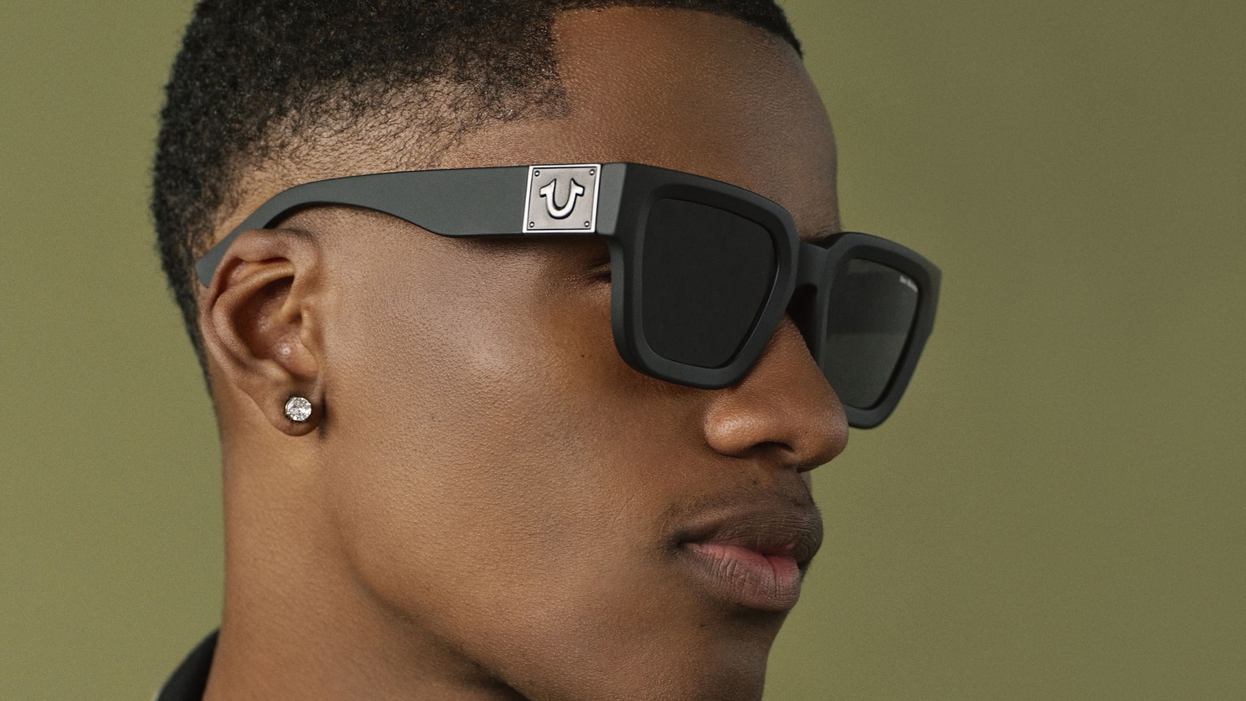 Close-up profile of a man wearing black square-frame True Religion sunglasses featuring the brand's horseshoe logo on the temple.