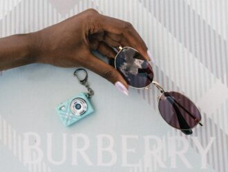 Burberry Spring Summer 2026 eyewear campaign — model holding round sunglasses and a mint check keychain camera against the iconic Burberry Check backdrop