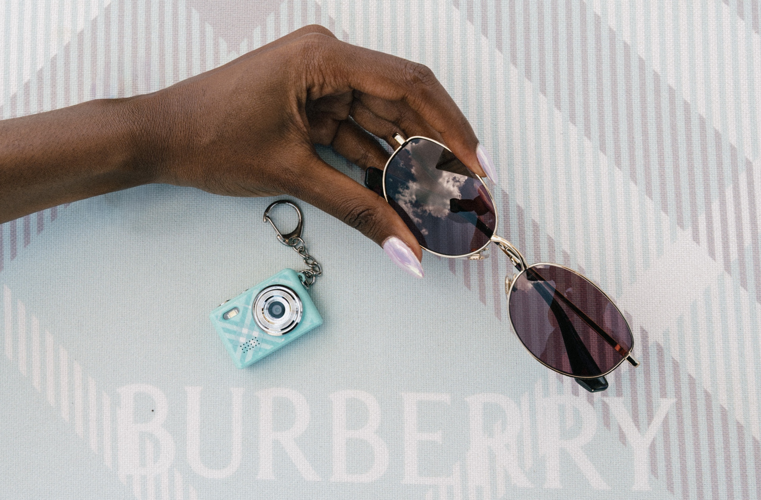 Burberry Spring Summer 2026 eyewear campaign — model holding round sunglasses and a mint check keychain camera against the iconic Burberry Check backdrop