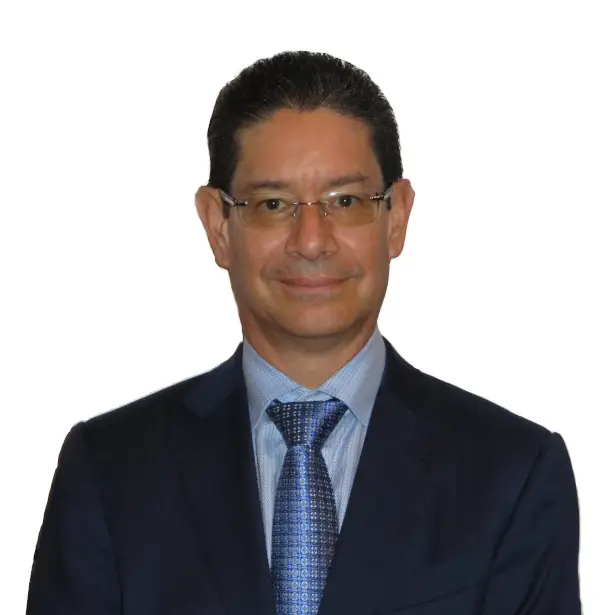 Portrait en plan rapproché du Dr Javier Prada-López, un homme souriant aux cheveux courts et sombres portant des lunettes de vue rimless, un costume bleu marine, une chemise bleu clair et une cravate bleue à motifs géométriques, sur fond blanc.