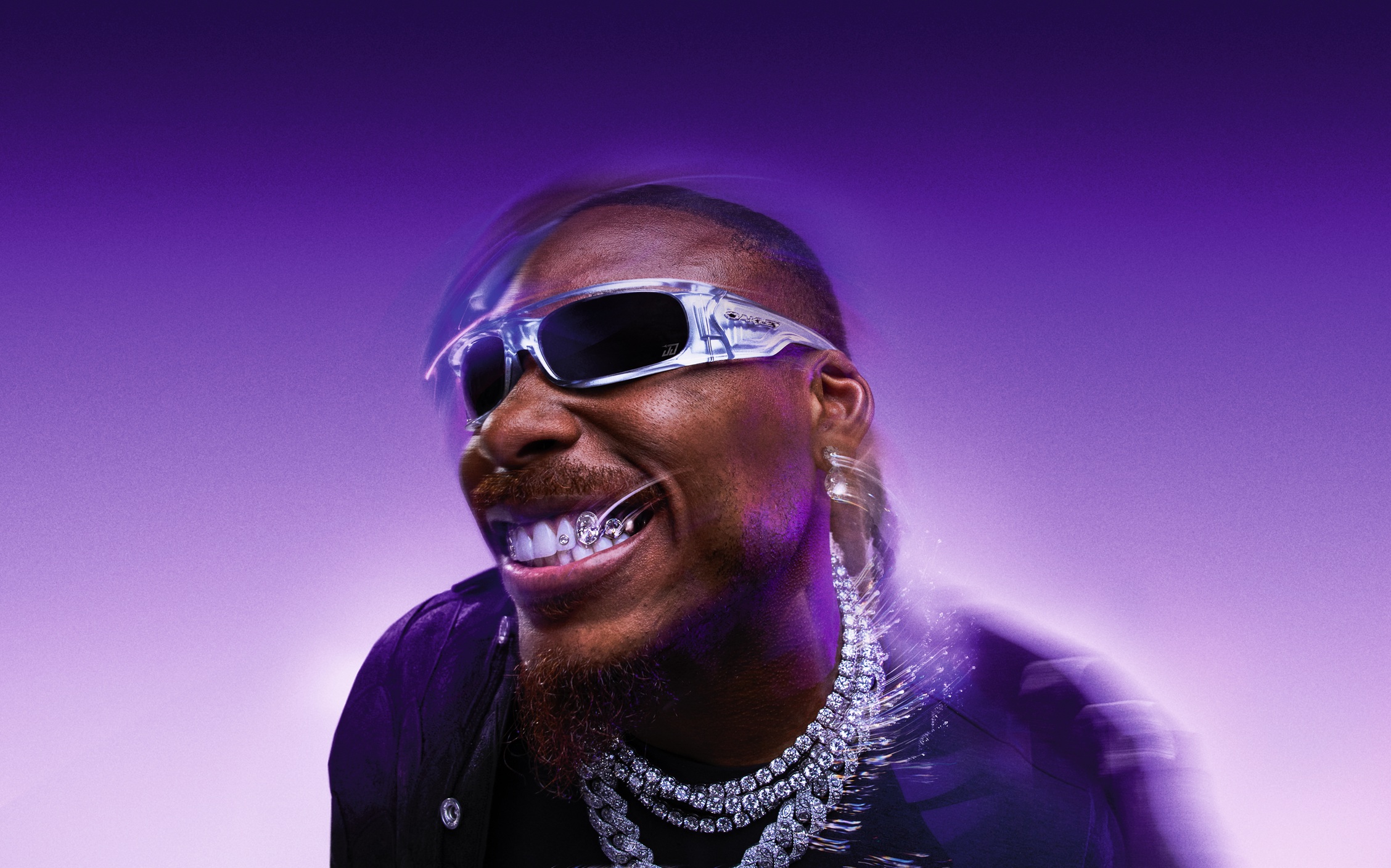 Justin Jefferson smiling in clear silver glass-effect Oakley Highland sunglasses with Prizm Black lenses and chrome detailing against a purple backdrop.