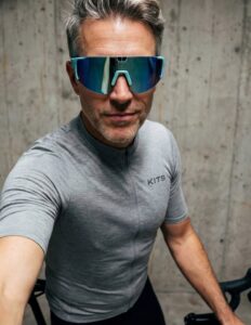 Un homme portant un maillot de cyclisme gris KITS et des lunettes connectées de cyclisme Pangolin 3 à verres réfléchissants bleus.