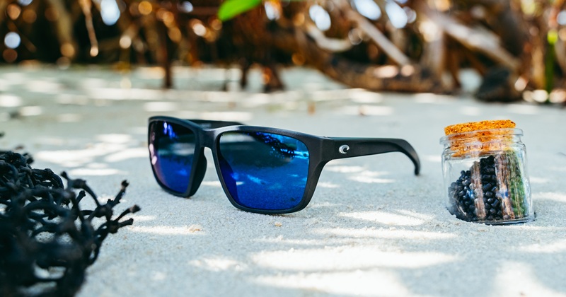 Costa Del Mar Rizzler black sunglasses with blue mirrored lenses on a sandy beach next to recycled NetPlus plastic pellets and fishing net.