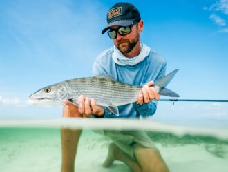 Angler in a cap and fishing shirt holding a caught bonefish in clear shallow water wearing Costa San Carlos sunglasses.