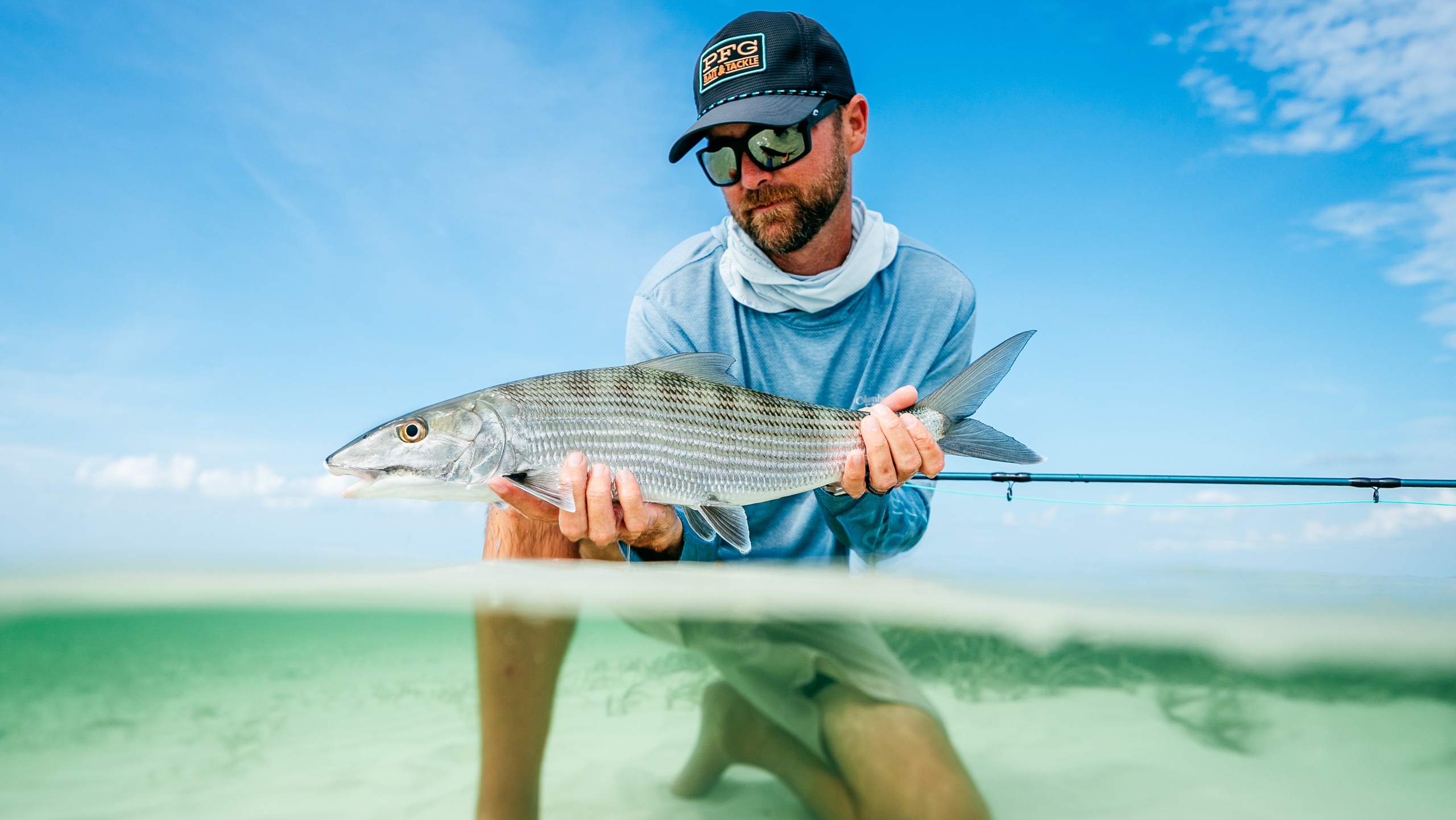 Angler in a cap and fishing shirt holding a caught bonefish in clear shallow water wearing Costa San Carlos sunglasses.