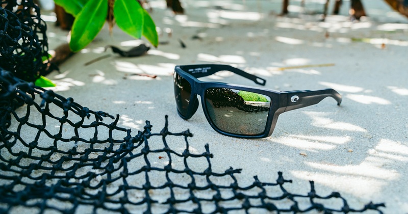 Costa Del Mar San Carlos matte black performance sunglasses resting on sand next to a black recycled fishing net.