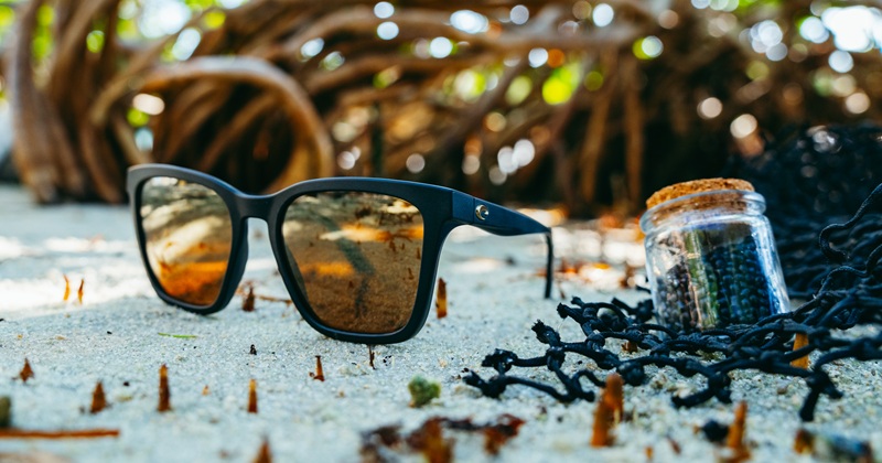 Costa Del Mar Sunlace women's matte black sunglasses with amber lenses on a sandy shore beside NetPlus recycled pellets.