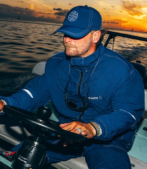 Homme pilotant un bateau au coucher du soleil, portant la veste bleue Costa Untangled Oceans, la casquette assortie et des lunettes de soleil autour du cou.