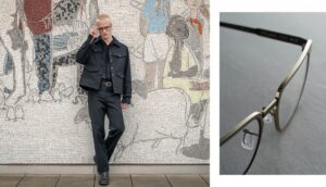 A male model dressed in black poses in front of an urban mosaic in Berlin, wearing Blackfin matte titanium architect-style eyeglass frames.