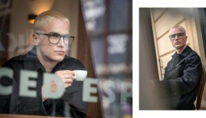 A man with short blond hair wearing luxury Blackfin prescription glasses, holding a cup through the window of an urban café.