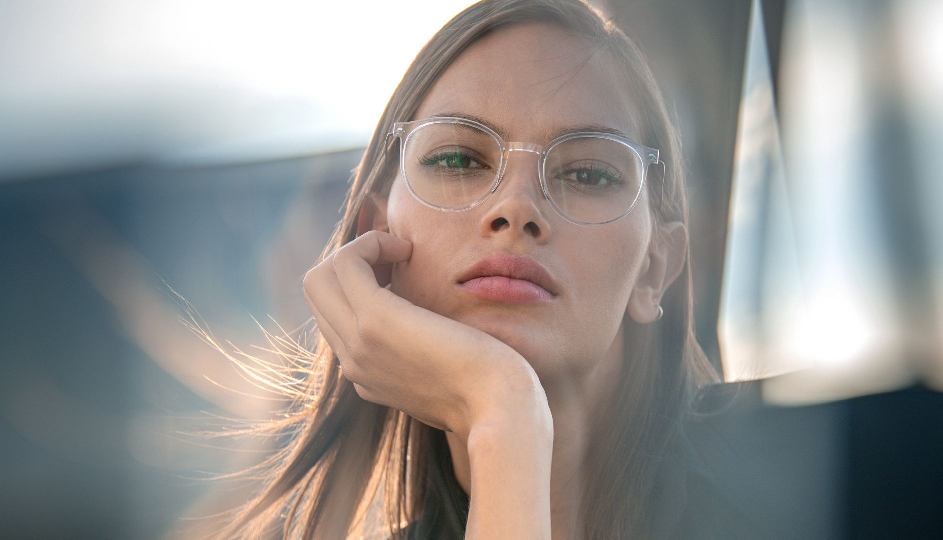 Gros plan d'un mannequin femme la tête posée sur sa main, portant des lunettes de vue Blackfin en titane transparentes sur fond urbain lumineux pour la campagne FW26 Urban Sync.
