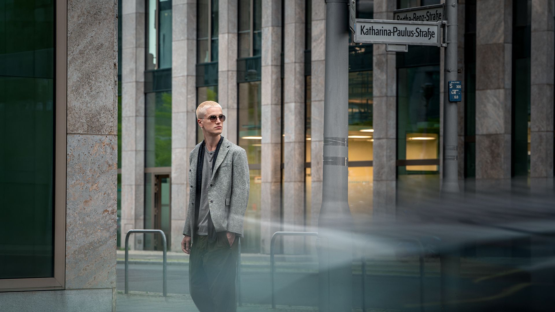 Full-body shot of a blonde male model wearing sunglasses and a blazer, standing near modern architecture on Katharina-Paulus-Straße in Berlin for the Blackfin FW26 Urban Sync campaign.