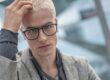 Portrait of a blonde male model running a hand through his hair, wearing dark, angular Blackfin titanium prescription glasses in an urban Berlin setting for FW26 Urban Sync.