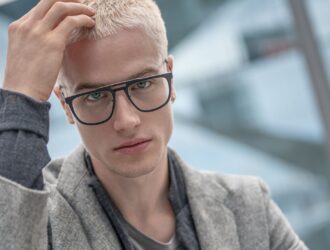 Portrait of a blonde male model running a hand through his hair, wearing dark, angular Blackfin titanium prescription glasses in an urban Berlin setting for FW26 Urban Sync.