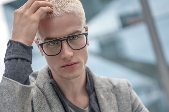 Portrait of a blonde male model running a hand through his hair, wearing dark, angular Blackfin titanium prescription glasses in an urban Berlin setting for FW26 Urban Sync.