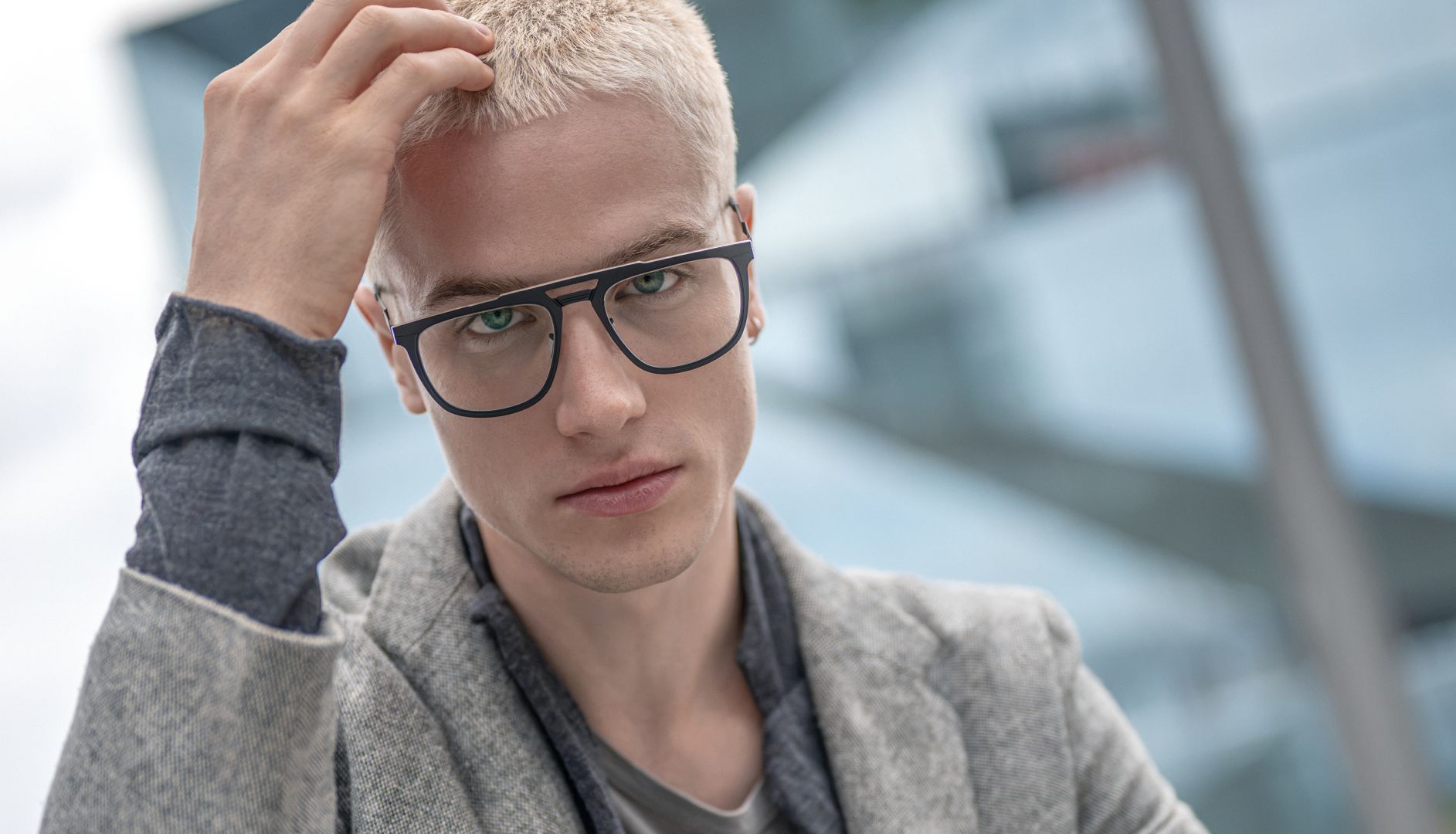 Portrait of a blonde male model running a hand through his hair, wearing dark, angular Blackfin titanium prescription glasses in an urban Berlin setting for FW26 Urban Sync.