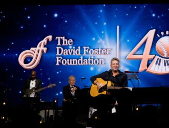Le musicien Chad Kroeger joue de la guitare acoustique sur scène devant un écran bleu affichant le logo du 40ème anniversaire de la David Foster Foundation.