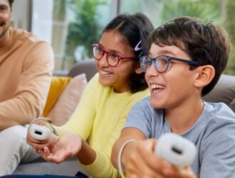 Two children wearing DOT myopia control spectacle lenses smiling and holding video game controllers while gaming comfortably at home.