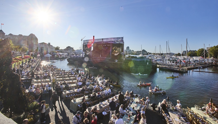 An outdoor, daytime view of an event setup along a harbor with long dining tables and a stage setup near docked boats.