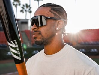 Vue de profil d'un joueur de baseball à l'extérieur tenant une batte Marucci, portant des lunettes masque Oakley noires en plein soleil.