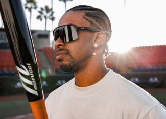 Profile view of a male baseball player outdoors holding a Marucci bat while wearing black Oakley shield sunglasses in bright sunlight.
