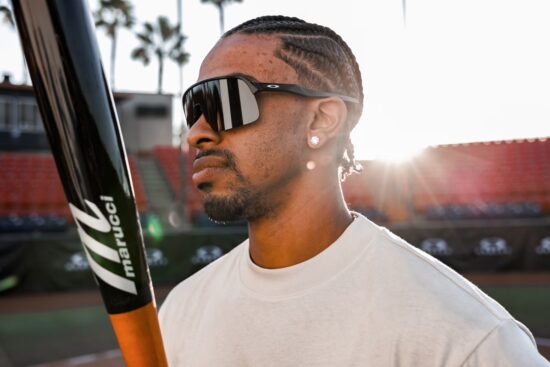 Profile view of a male baseball player outdoors holding a Marucci bat while wearing black Oakley shield sunglasses in bright sunlight.
