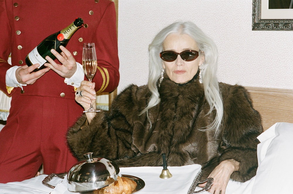 Senior woman wearing LINDA FARROW oval sunglasses and a fur coat receiving room service champagne in a luxury hotel bed.