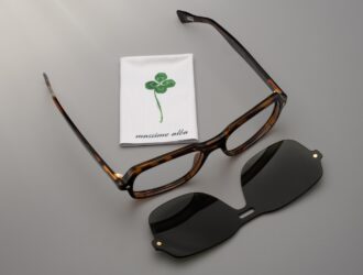 Monture de lunettes en acétate écaille de tortue signée SNOB Milano x Massimo Alba, posée sur un fond neutre à côté d'un clip solaire magnétique noir et d'une suédine blanche marquée du logo trèfle à quatre feuilles vert.