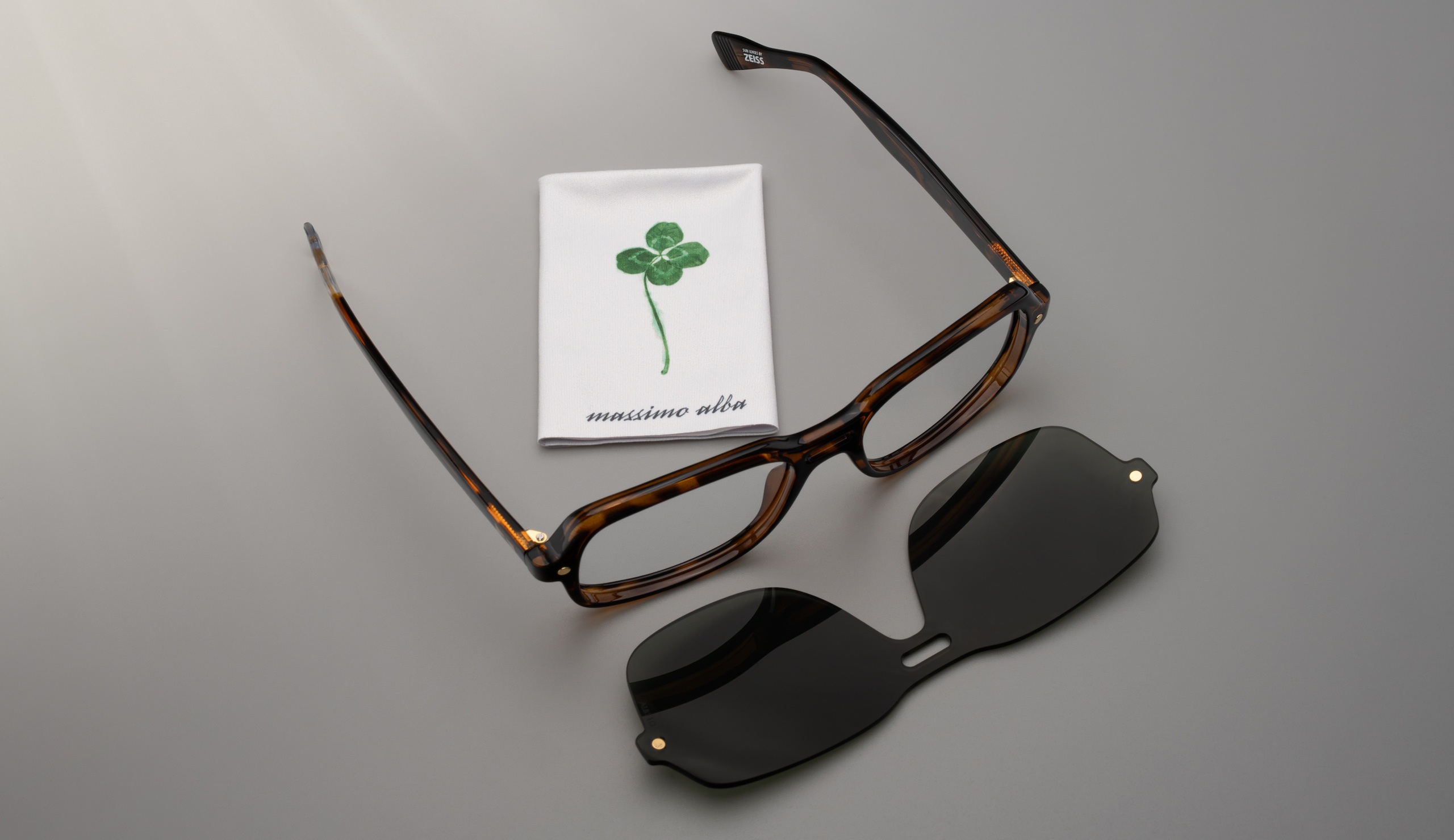 Monture de lunettes en acétate écaille de tortue signée SNOB Milano x Massimo Alba, posée sur un fond neutre à côté d'un clip solaire magnétique noir et d'une suédine blanche marquée du logo trèfle à quatre feuilles vert.