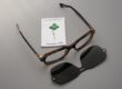 Tortoiseshell acetate eyewear frame by SNOB Milano x Massimo Alba resting on a neutral background alongside a black magnetic clip-on sun lens and a branded white microfiber cleaning cloth featuring a green four-leaf clover logo.