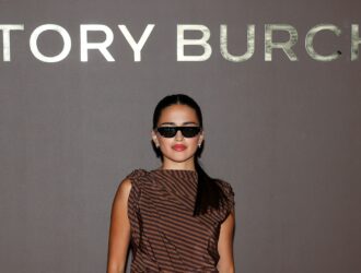 Maia Reficco pose devant le mur photo de Tory Burch, portant des lunettes de soleil noires épurées et un haut à rayures brunes à une épaule, cheveux foncés attachés en queue de cheval basse.
