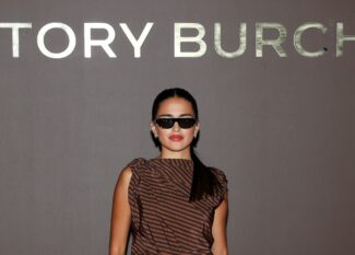 Maia Reficco poses in front of the Tory Burch step-and-repeat wall wearing sleek black sunglasses and a brown striped one-shoulder top, her dark hair pulled into a low ponytail.