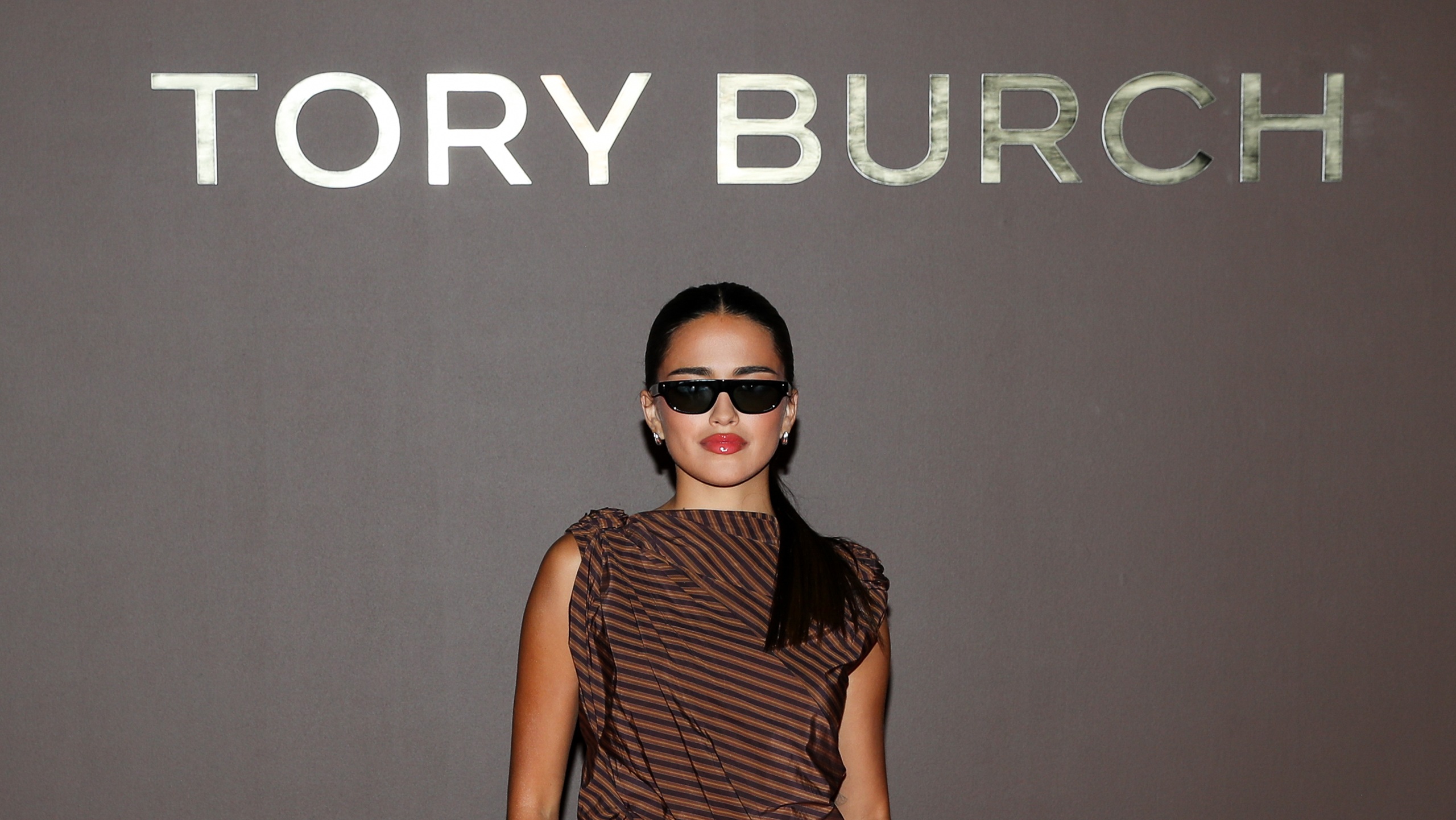 Maia Reficco poses in front of the Tory Burch step-and-repeat wall wearing sleek black sunglasses and a brown striped one-shoulder top, her dark hair pulled into a low ponytail.
