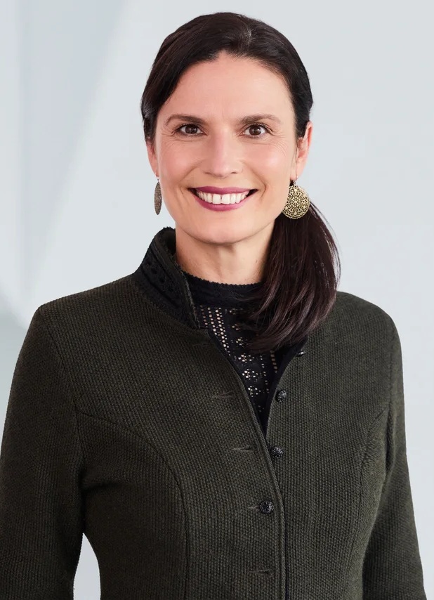 Portrait of Cathleen Kabashi, Exhibition Director of opti, smiling in a green jacket with dark hair pulled back into a side ponytail.