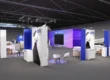 Preliminary 3D rendering of the opti SMART TECH exhibition area, featuring modern blue and white display stands, seating areas, and a purple background wall labeled "opti SMART EYEWEAR.