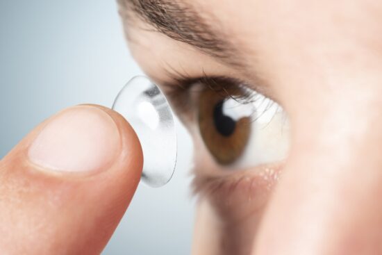 Close-up of a person using a fingertip to insert a clear contact lens into their brown eye.