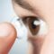 Close-up of a person using a fingertip to insert a clear contact lens into their brown eye.