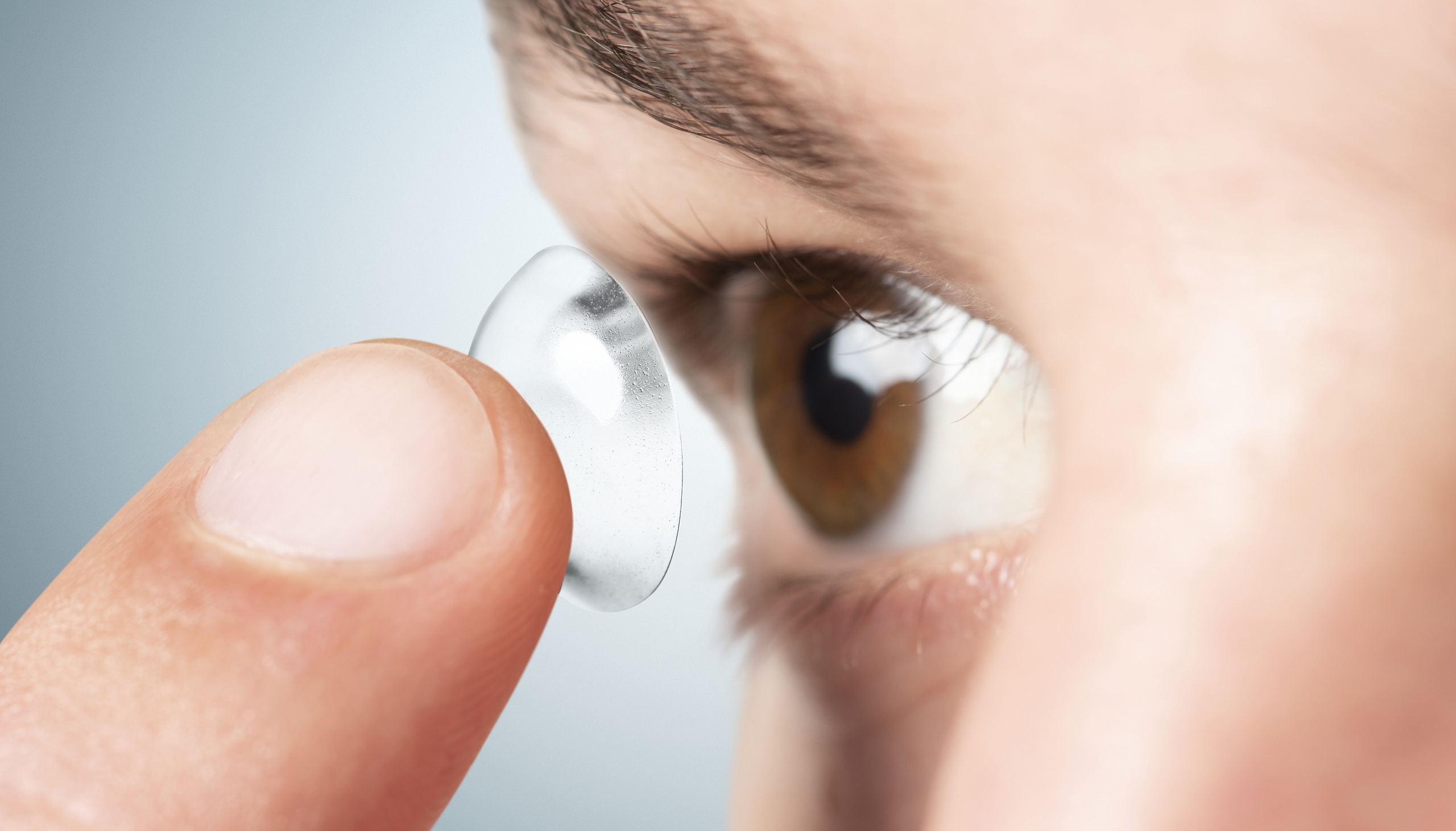 Close-up of a person using a fingertip to insert a clear contact lens into their brown eye.