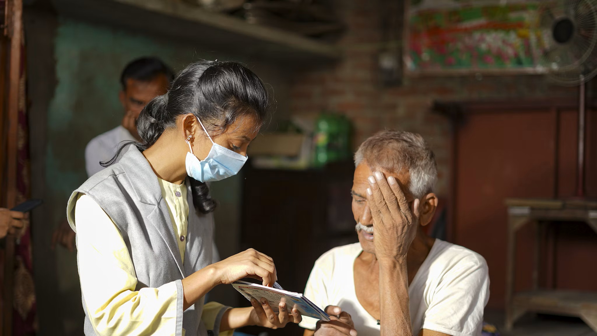 An eye care worker wearing a face mask conducts a vision screening for an elderly man covering one eye in India.