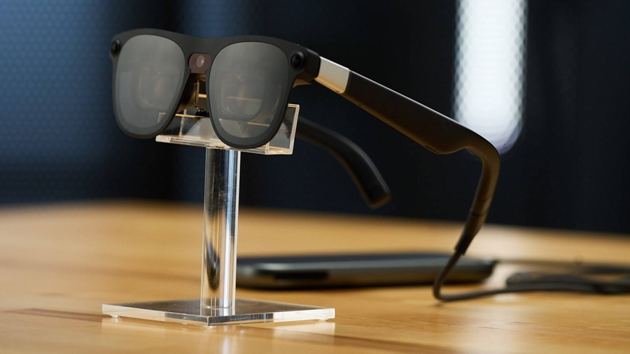XREAL AURA AR smart glasses displayed on an acrylic stand on a wooden surface with a connected compute puck in the background.