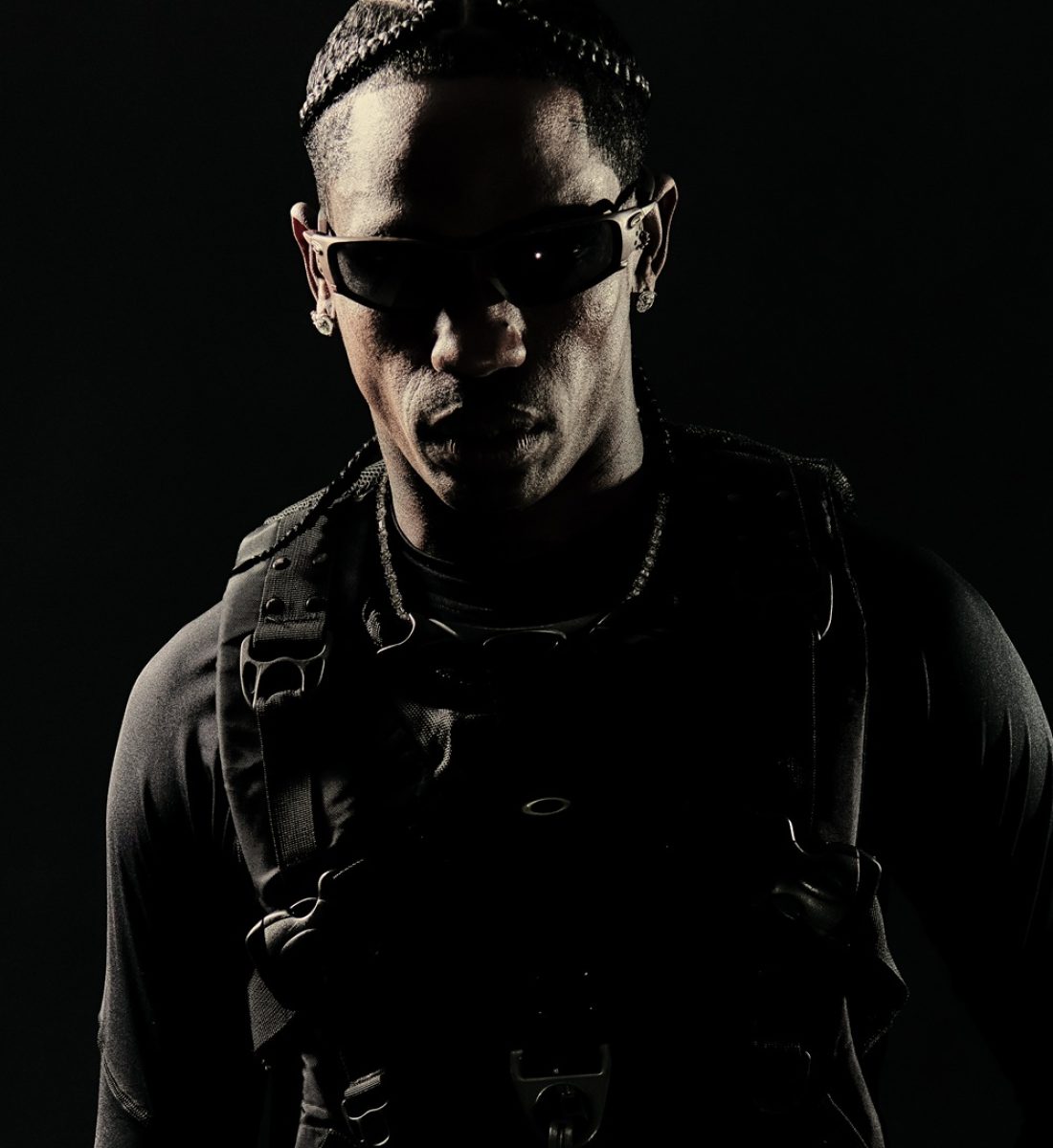 Travis Scott wearing Oakley sunglasses in a dramatic black and white portrait, dressed in dark tactical-style clothing with straps and buckles against a black background, showcasing the post-apocalyptic aesthetic from his collaboration with Oakley